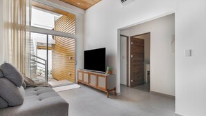 Ein modernes Wohnzimmer mit einer grauen Couch, einem Holz-TV-Schrank und einem großen Fenster mit Jalousien.
