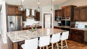 A modern kitchen with dark wooden cabinets, stainless steel appliances, a granite island with bar stools, and pendant lighting. - Luxurious Lakefront Escape – Premier EAA Rental with Breathtaking Views (Winneconne)