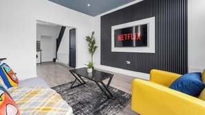Un salon moderne aux couleurs noir et blanc, avec un canapé jaune, une table basse noire et un téléviseur fixé au mur qui affiche le logo « NETFLIX ».