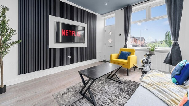 Un salon moderne avec un grand téléviseur à écran plat fixé sur un mur noir, un fauteuil jaune, une table basse noire et une vue sur l’extérieur par le biais d’une grande fenêtre.