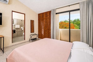 A hotel room with a bed, a small table, a mirror, a wardrobe, and a balcony with a view of trees. - Breezy Studio in Cancún with Balcony & Park View (Cancún)