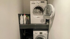 Une petite buanderie comprenant deux machines à laver superposées, une étagère avec des bouteilles de lessive et une poubelle.