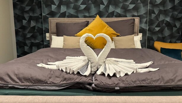 Un lit avec une tête de lit ornée d’un motif géométrique, une serviette décorative en forme de cœur et un coussin jaune.