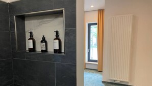 Une salle de bain avec des carreaux de couleur foncée, une étagère avec trois bouteilles, un radiateur et une fenêtre avec des rideaux.
