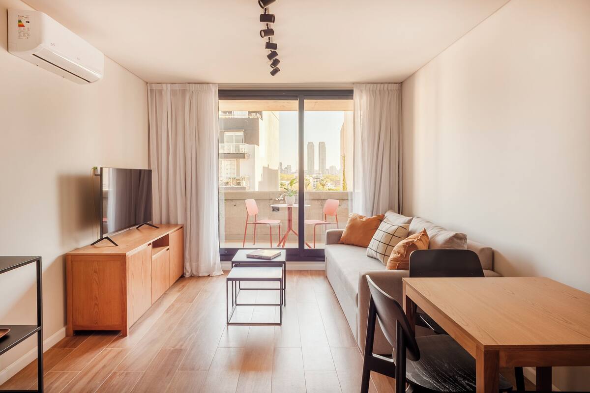 Una sala de estar moderna con sofá, una mesa de comedor, televisor y un balcón con sillas.