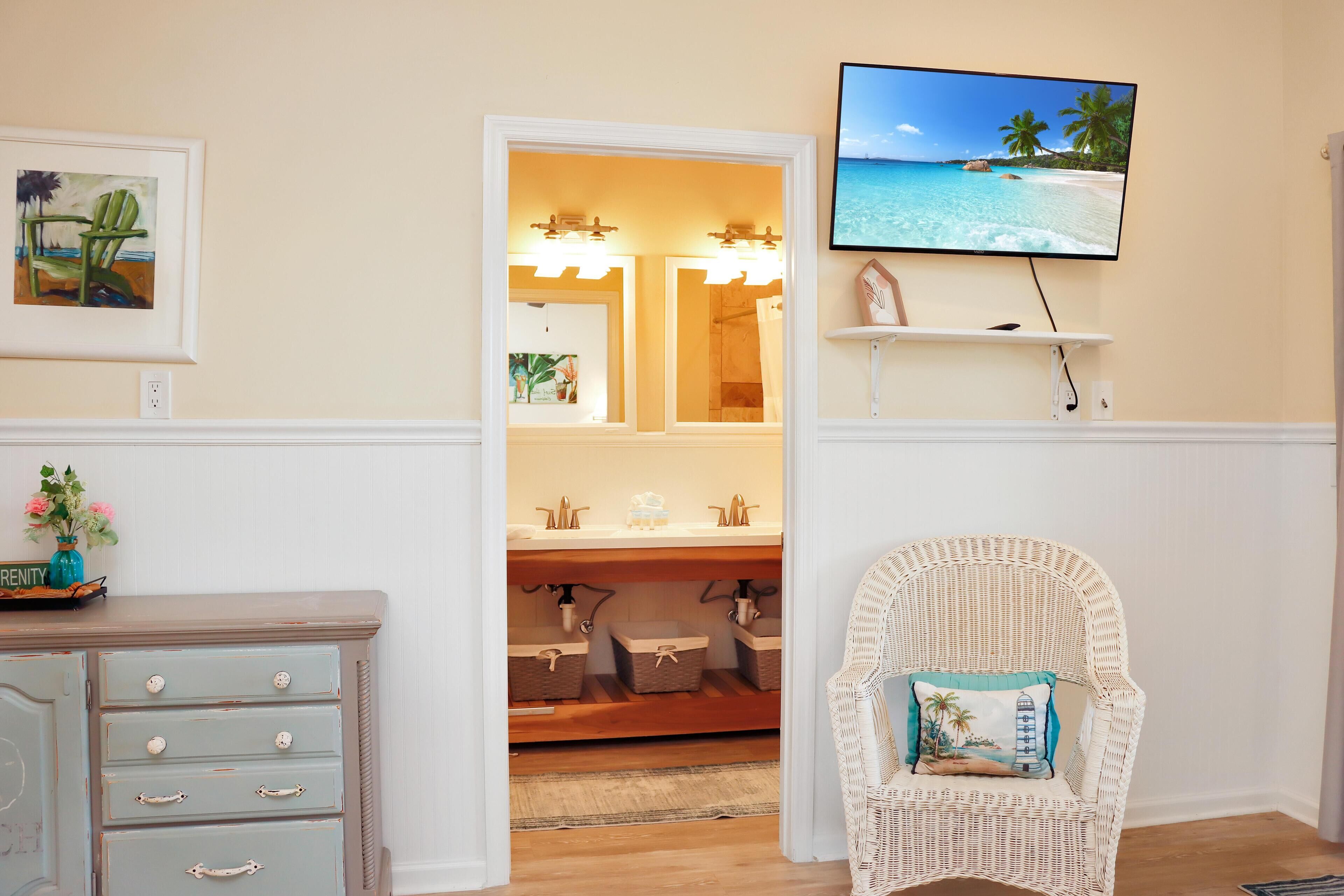 A bathroom with a vanity, a mirror, a television mounted above the sink, and a wicker chair with a cushion.