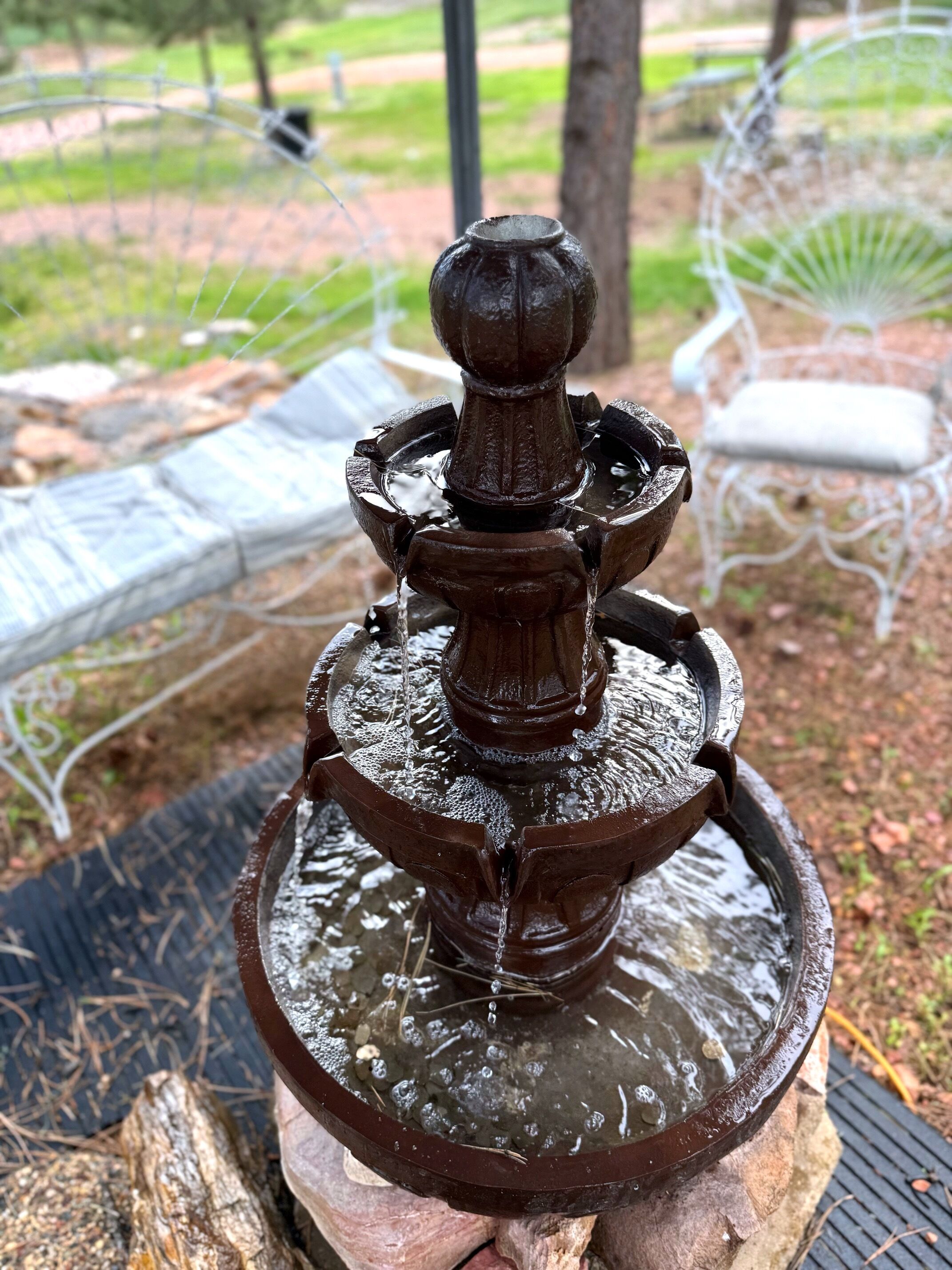 Une fontaine en métal, dont l’eau jaillit de l’étage supérieur, est posée sur un socle en pierre dans un espace extérieur. On aperçoit également une chaise en métal et un arbre en arrière-plan.