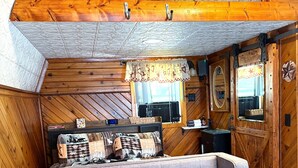 Un intérieur de chalet chaleureux avec un canapé gris à motifs capitonnés, des murs en bois, un plafond orné d’un motif décoratif, une horloge et une pancarte de bienvenue.