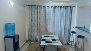 A compact living space with a dining table, two bar stools, a water dispenser, and a small kitchenette area.