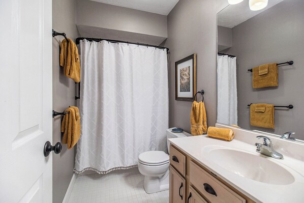 Un baño con cortina de ducha blanca, un inodoro, un lavabo con espejo y toallas colgadas en la pared.