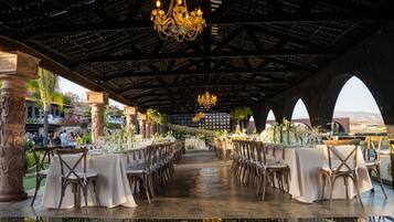 An indoor venue with a long table set for dining, chandeliers, and a view of the outdoors.