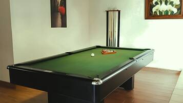 A pool table with a green felt surface, a cue ball, and a red object on the table. There is a framed picture of flowers on the wall to the right.