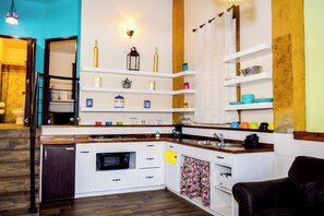 Une cuisine compacte avec des meubles blancs, un four à micro-ondes, un évier et une cuisinière. Des étagères au-dessus du plan de travail servent à ranger divers ustensiles de cuisine.
