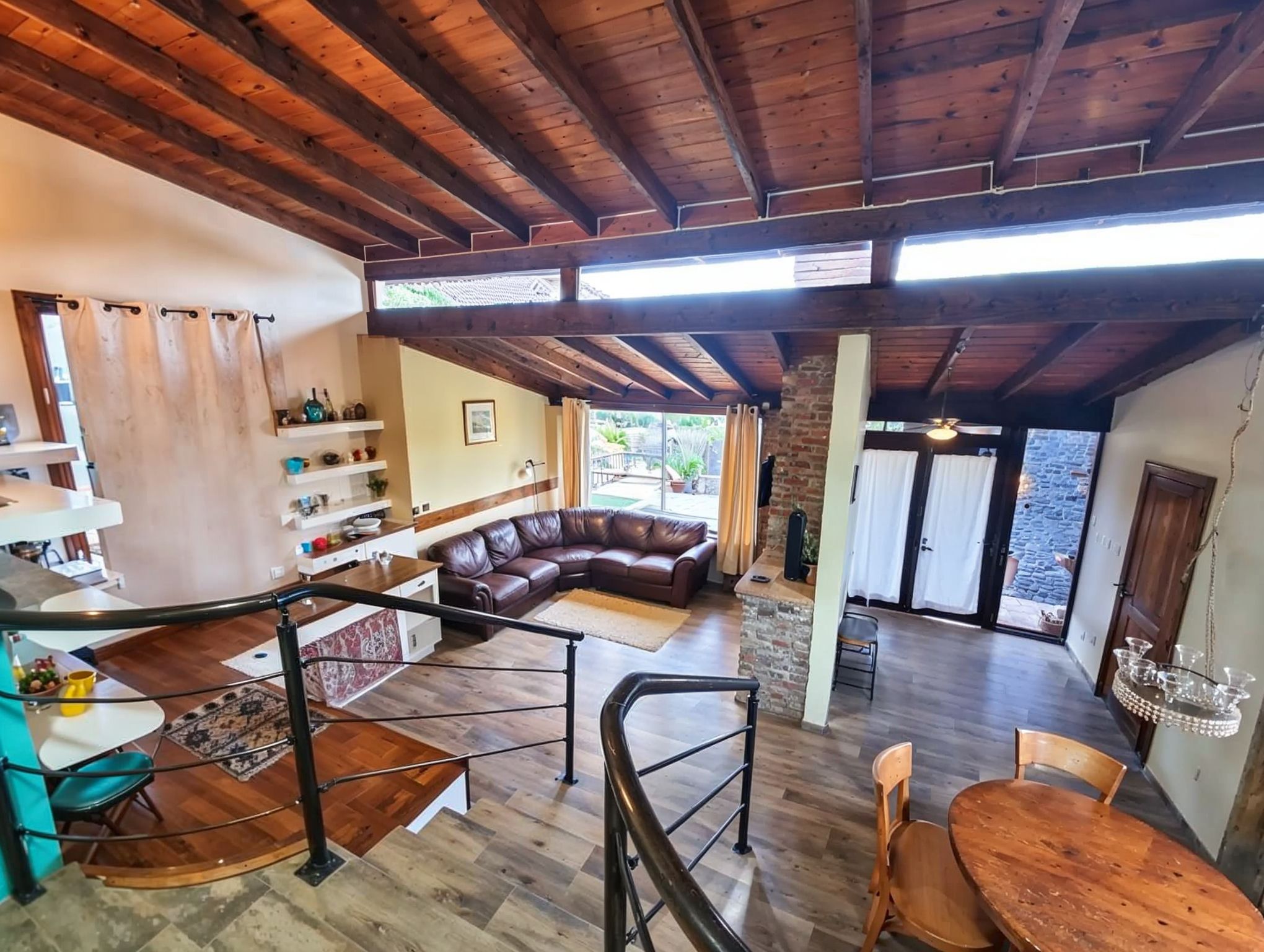 A spacious living area with a wooden ceiling, a brown leather sofa, a dining table with chairs, and a staircase with a railing.