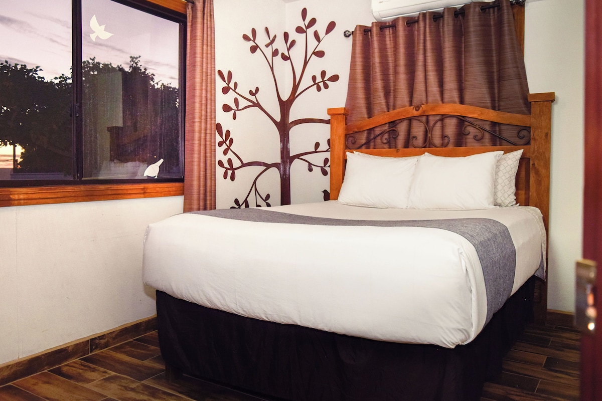 A hotel room with a bed, a window with curtains, a wall decoration, and an air conditioning unit.