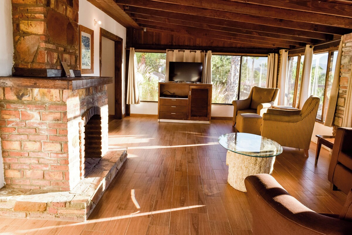 A spacious living room with a brick fireplace, wooden flooring, a television, and a comfortable seating area with a glass coffee table.