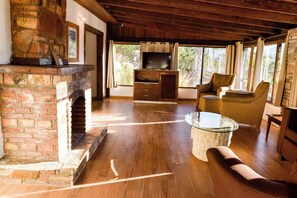 A spacious living room with a brick fireplace, wooden flooring, a glass coffee table, and wicker furniture. - Finca Selah (Valle de Guadalupe)