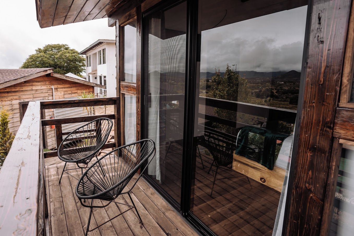 Un balcón con suelo de madera, dos sillas negras de metal y un ventanal con vistas a un paisaje urbano.