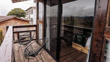 Un balcón con suelo de madera, dos sillas negras de metal y un ventanal con vistas a un paisaje urbano.