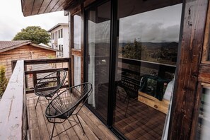 A balcony with a wooden floor, two black wire chairs, and a glass door leading to a view of a cityscape.