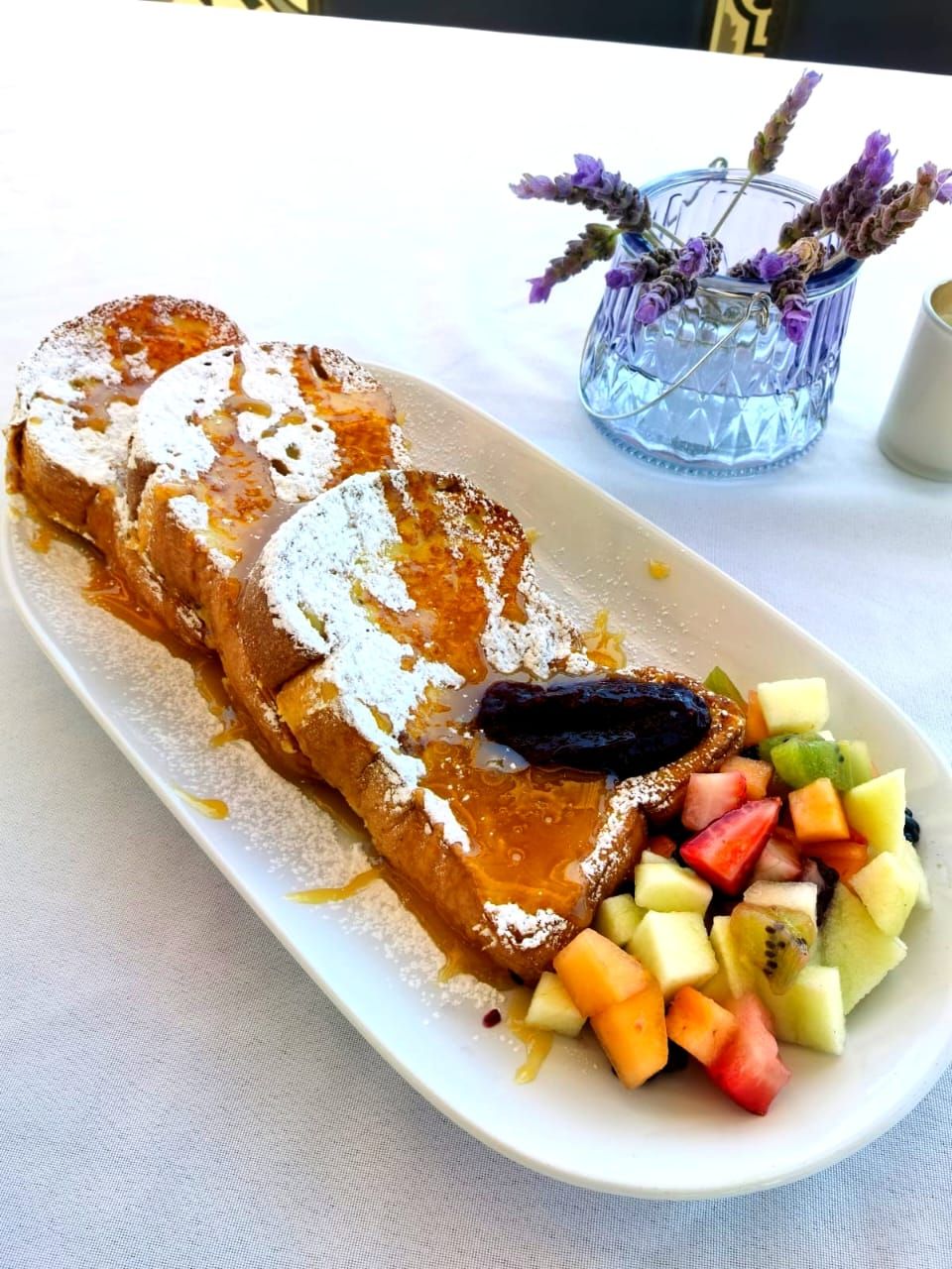 Un plato de desayuno con tostadas francesas, jarabe, azúcar glas y una ensalada de frutas.