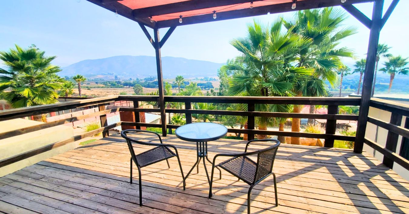 A wooden deck with two chairs and a table, overlooking a landscape with palm trees and mountains.