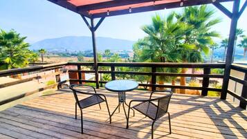 A wooden deck with two chairs and a table, overlooking a landscape with palm trees and mountains.