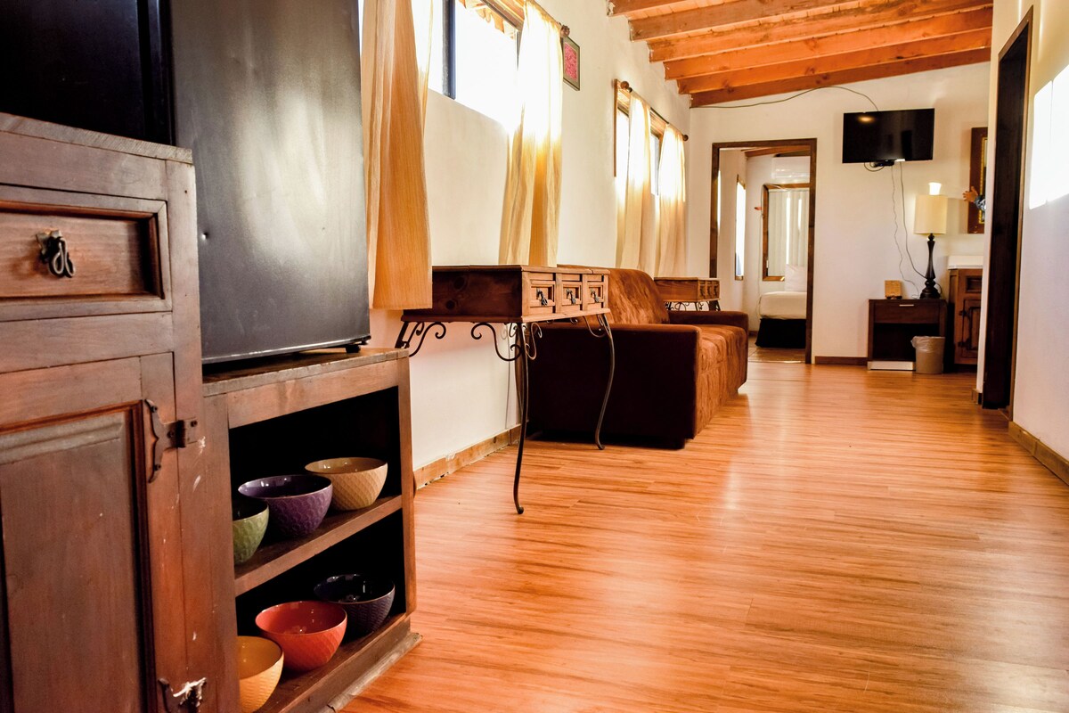 A spacious living area with wooden flooring, a sofa, a television, and a cabinet with bowls.