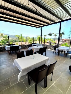An outdoor dining area with tables and chairs, a pergola overhead, and a view of a landscape with trees and hills.