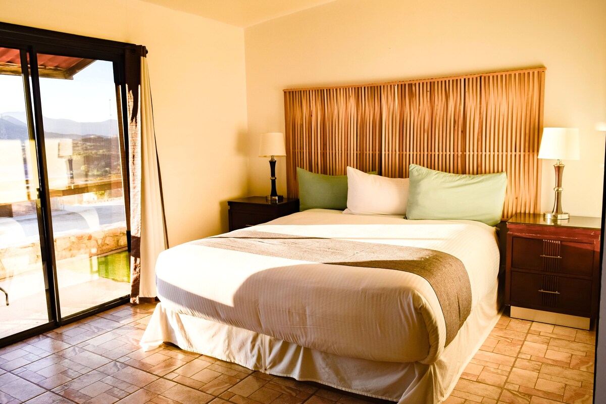 A hotel room with a bed, a nightstand, a lamp, a window with curtains, and a view of the outdoors.