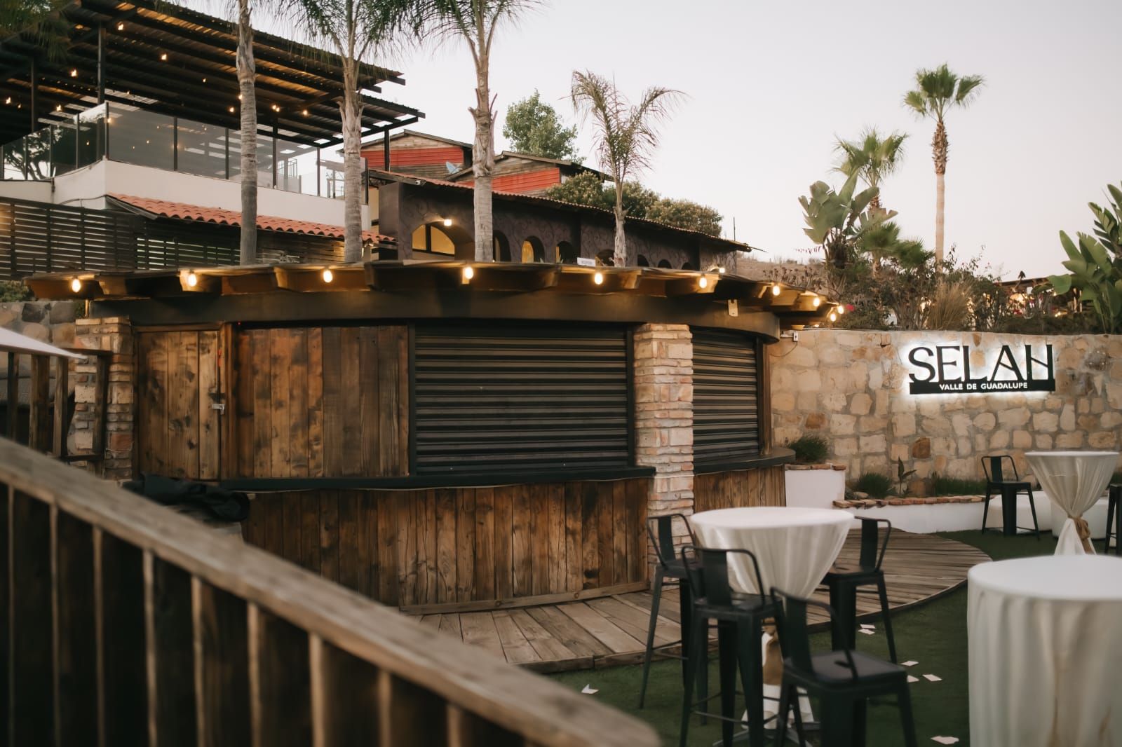 An outdoor seating area with wooden tables and chairs, a stone wall with the name 'SELAN', and a wooden bar with lights.