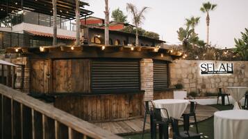 An outdoor seating area with wooden tables and chairs, a stone wall with the name 'SELAN', and a wooden bar with lights.