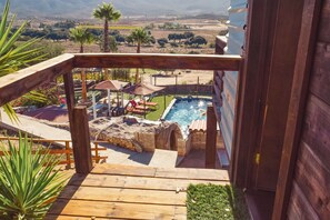 A wooden deck leading to a swimming pool with lounge chairs and umbrellas, surrounded by greenery and a view of distant hills.