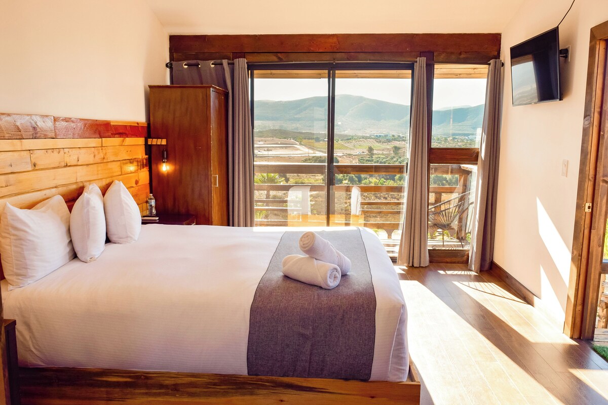 A bedroom with a large bed, a wooden headboard, a nightstand, a TV, and a view of a mountainous landscape through a sliding door.