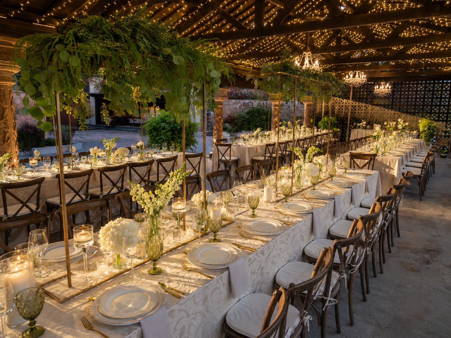 Un área de comedor al aire libre con mesas dispuestas para un evento formal, con manteles blancos, cubiertos dorados y centros de mesa con velas.