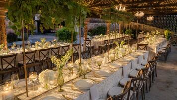 Un área de comedor al aire libre con mesas dispuestas para un evento formal, con manteles blancos, cubiertos dorados y centros de mesa con velas.