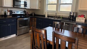 A modern kitchen with white and dark blue cabinets, stainless steel appliances, a wooden dining table, and a coffee bar.