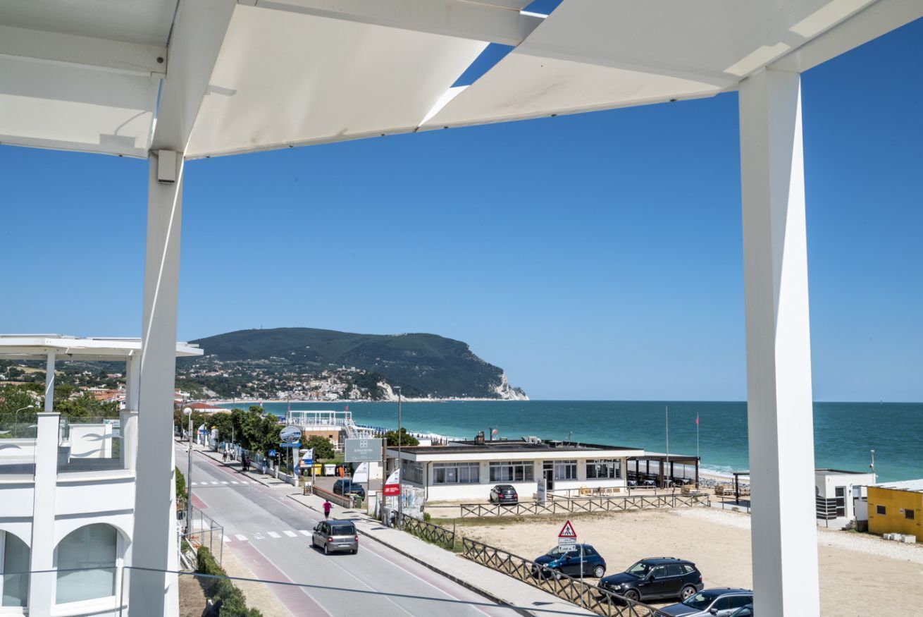 Une route côtière bordée d’un bâtiment situé au bord de la plage, un ciel bleu dégagé et une colline lointaine.