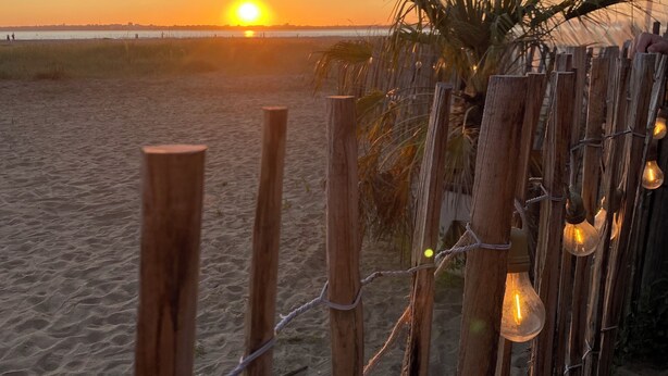 A beach scene with a wooden fence and string lights, a sunset in the background, and a palm tree.
