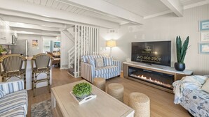 A modern living room with a fireplace, a television, and a dining area with wicker chairs. - 5BR/4BA-Oceanview-Pool-Sundeck-Bike-Theater-Game Room-Sleep 16+ (Ocean City)