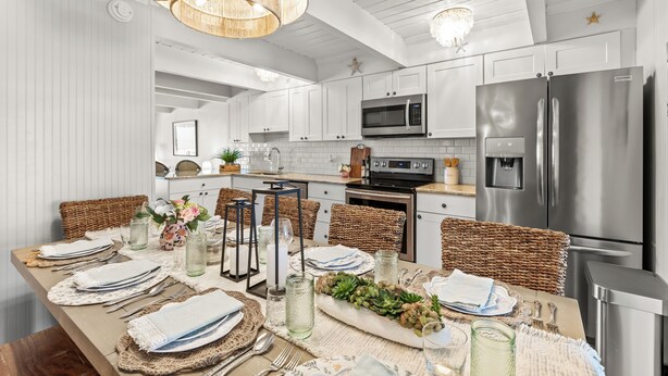 A modern kitchen with white cabinetry, stainless steel appliances, and a dining area set with wicker chairs and a wooden table. - 5BR/4BA-Oceanview-Pool-Sundeck-Bike-Theater-Game Room-Sleep 16+ (Ocean City)