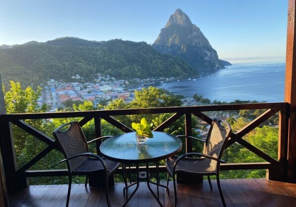 A balcony with a glass table and chairs, overlooking a coastal town and a prominent mountain.