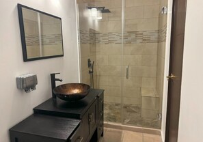 A bathroom with a large mirror, a black vanity with a bowl sink, a towel dispenser, and a glass-enclosed shower area.