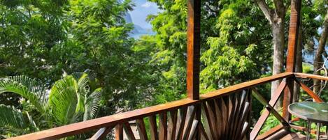 A wooden deck with a metal table, surrounded by lush green foliage and a mountain in the background.
