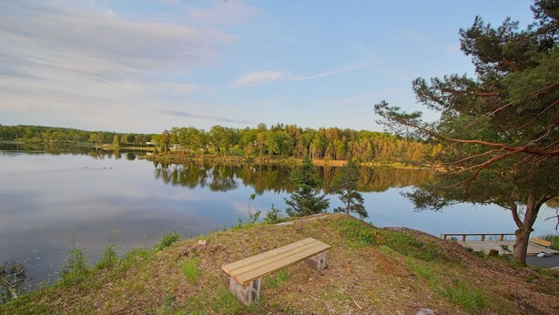 A bench by a calm lake with a forest in the background. - Ansgarsgården (Flen)