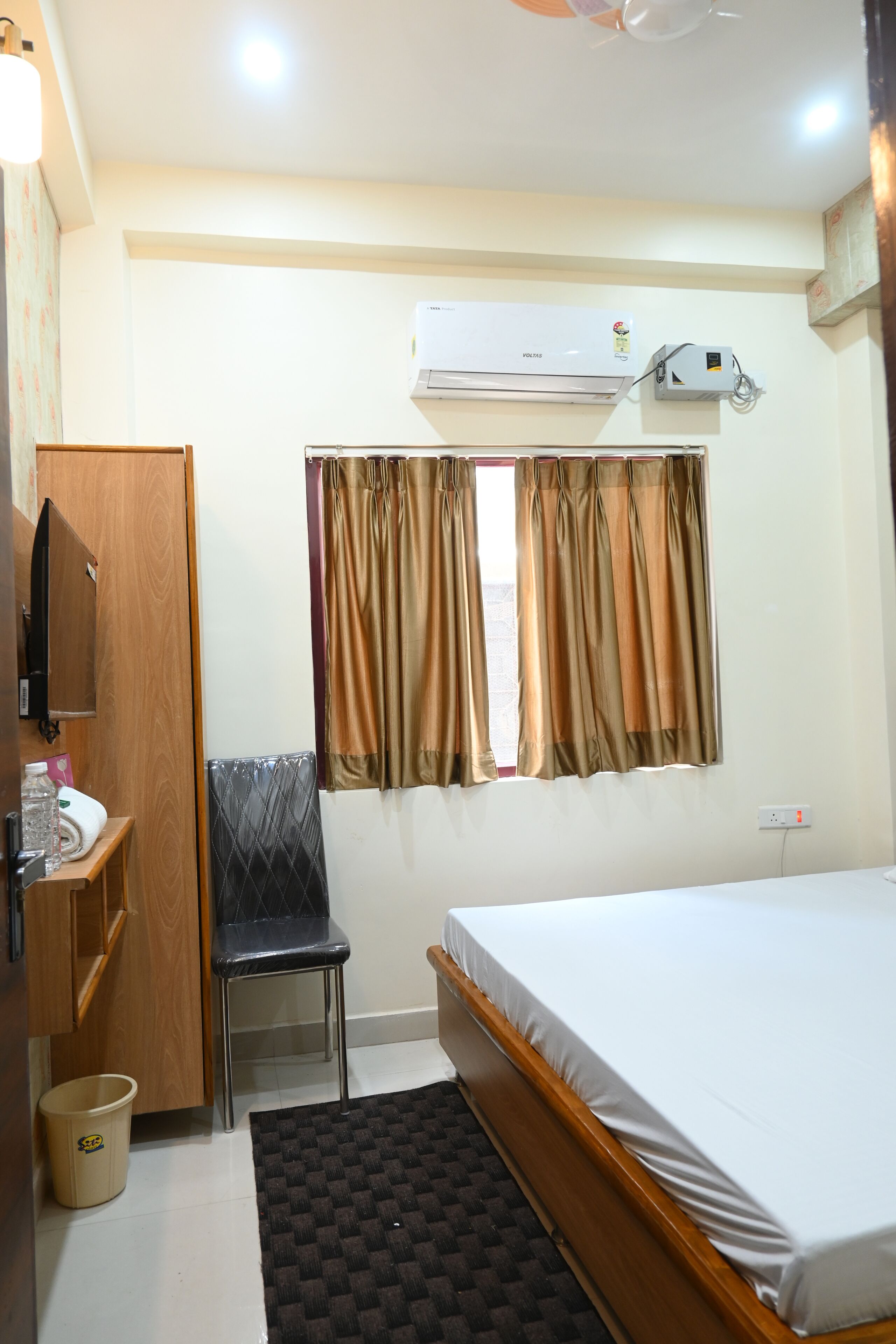 A hotel room with a bed, a chair, a small table, a wardrobe, an air conditioning unit, and a window with curtains.