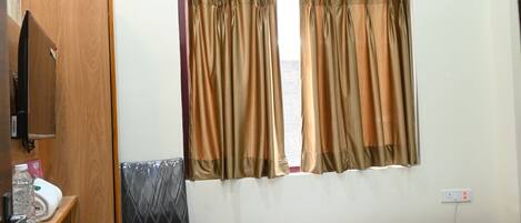 A hotel room with a bed, a chair, a small table, a wardrobe, an air conditioning unit, and a window with curtains.