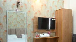 Habitación de hotel con cama, espejo, televisor, armario de madera, estante pequeño con artículos de aseo y un cubo de basura.