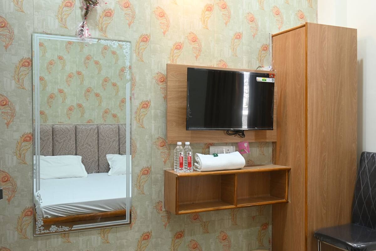 Quarto de hotel com cama, espelho, televisão, guarda-roupa de madeira, prateleira pequena com produtos de higiene e um cesto de lixo.