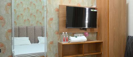 A hotel room with a bed, a mirror, a television, a wooden wardrobe, a small shelf with toiletries, and a trash bin.